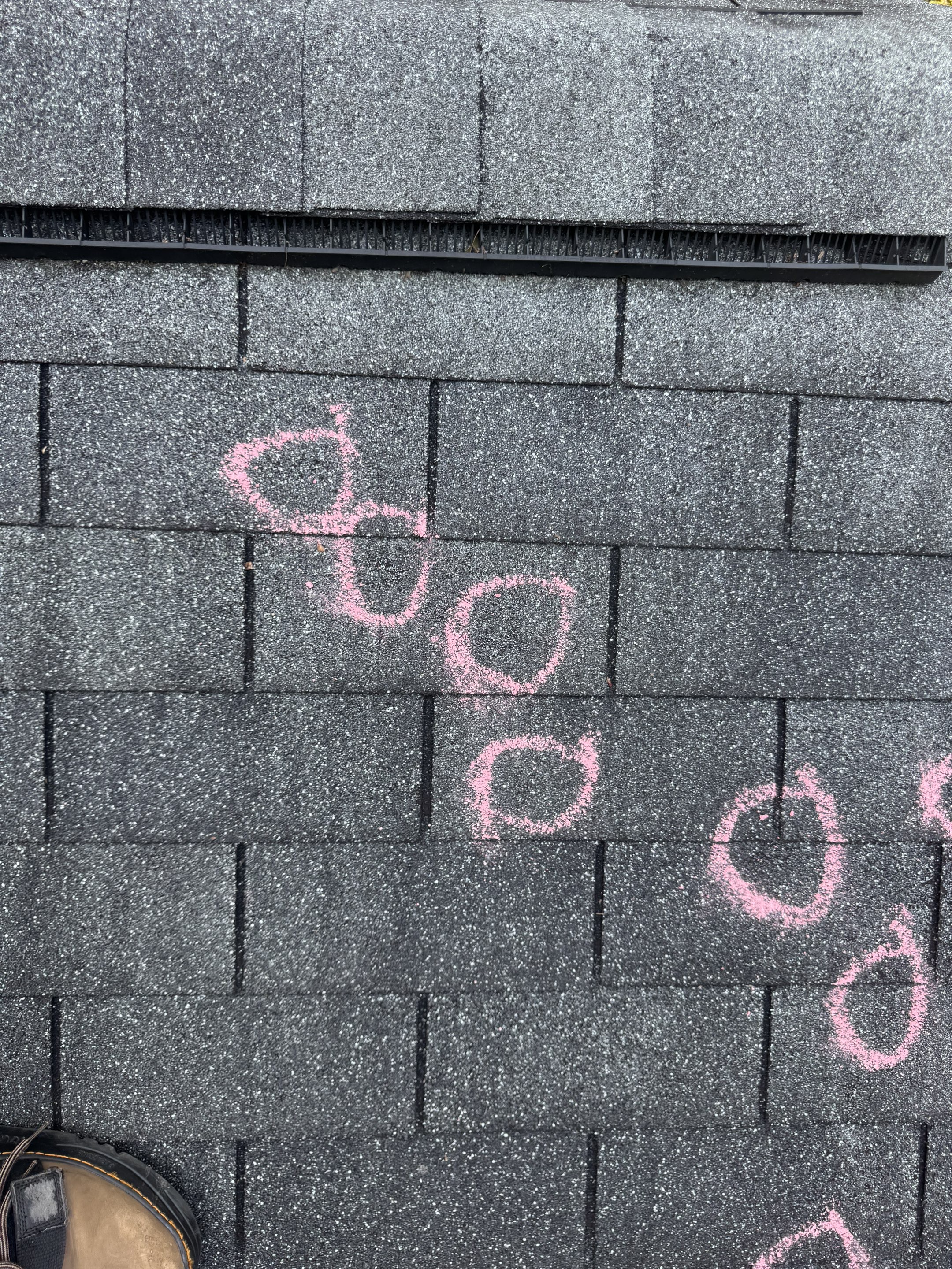 Close-up of dark asphalt shingles with pink chalk circles around individual hail strikes, documented for insurance claim